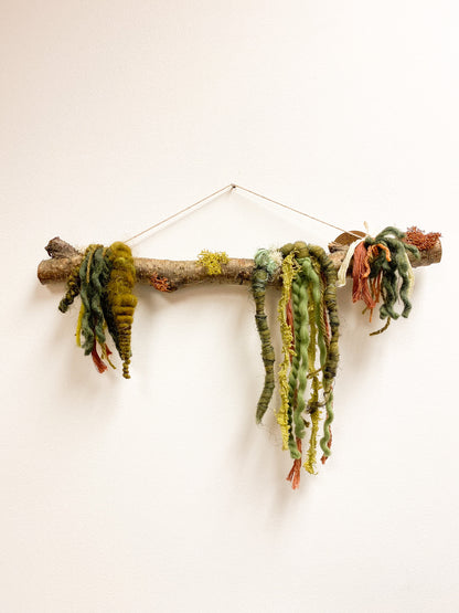 Decorative wall hanging made of a branch with knotted green and brown yarn on a white background