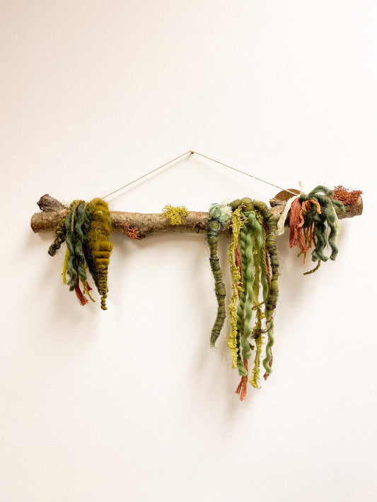 Decorative wall hanging made of a branch with knotted green and brown yarn on a white background