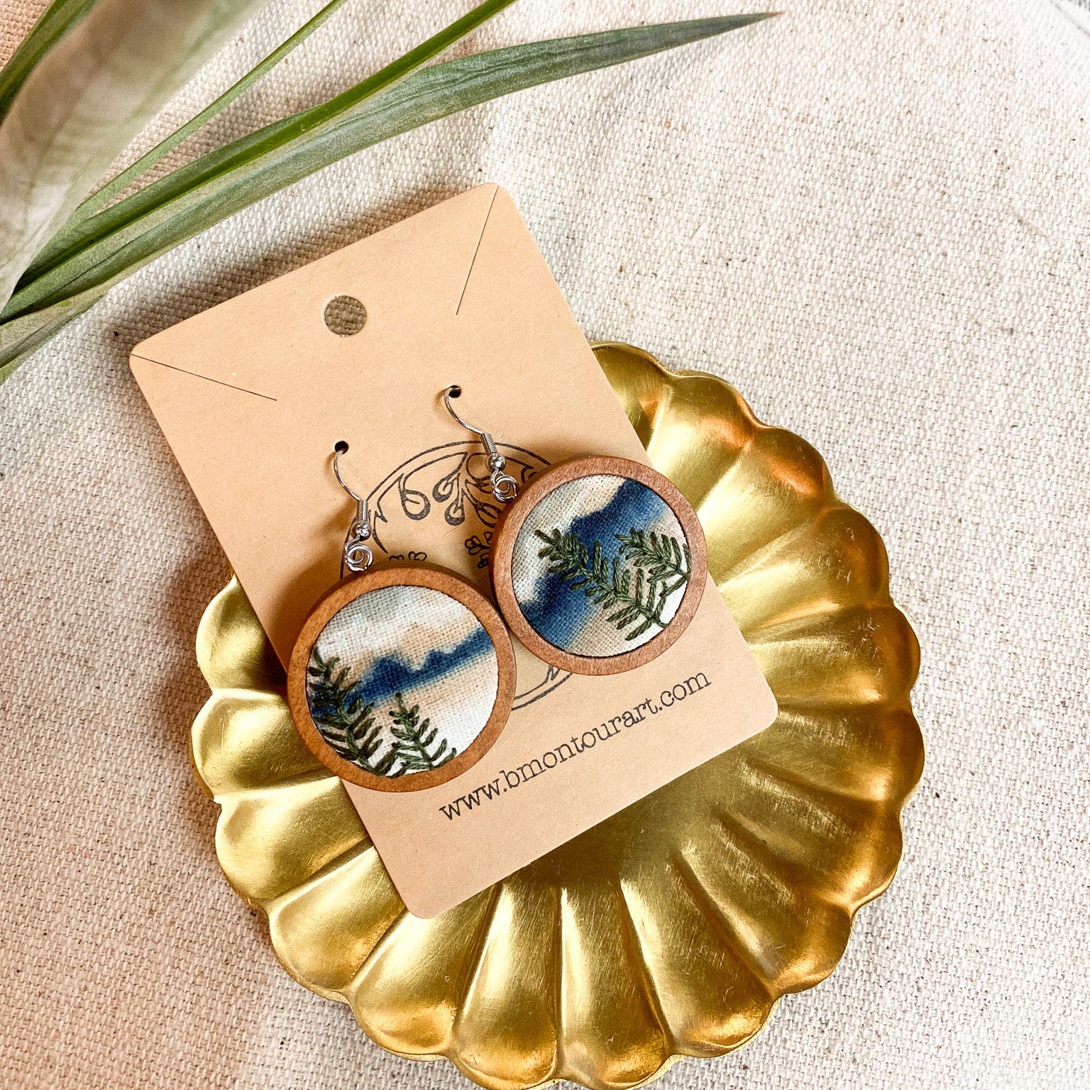 Pair of earrings on a gold display stand with a scenic design, placed on a textured surface with green leaves.