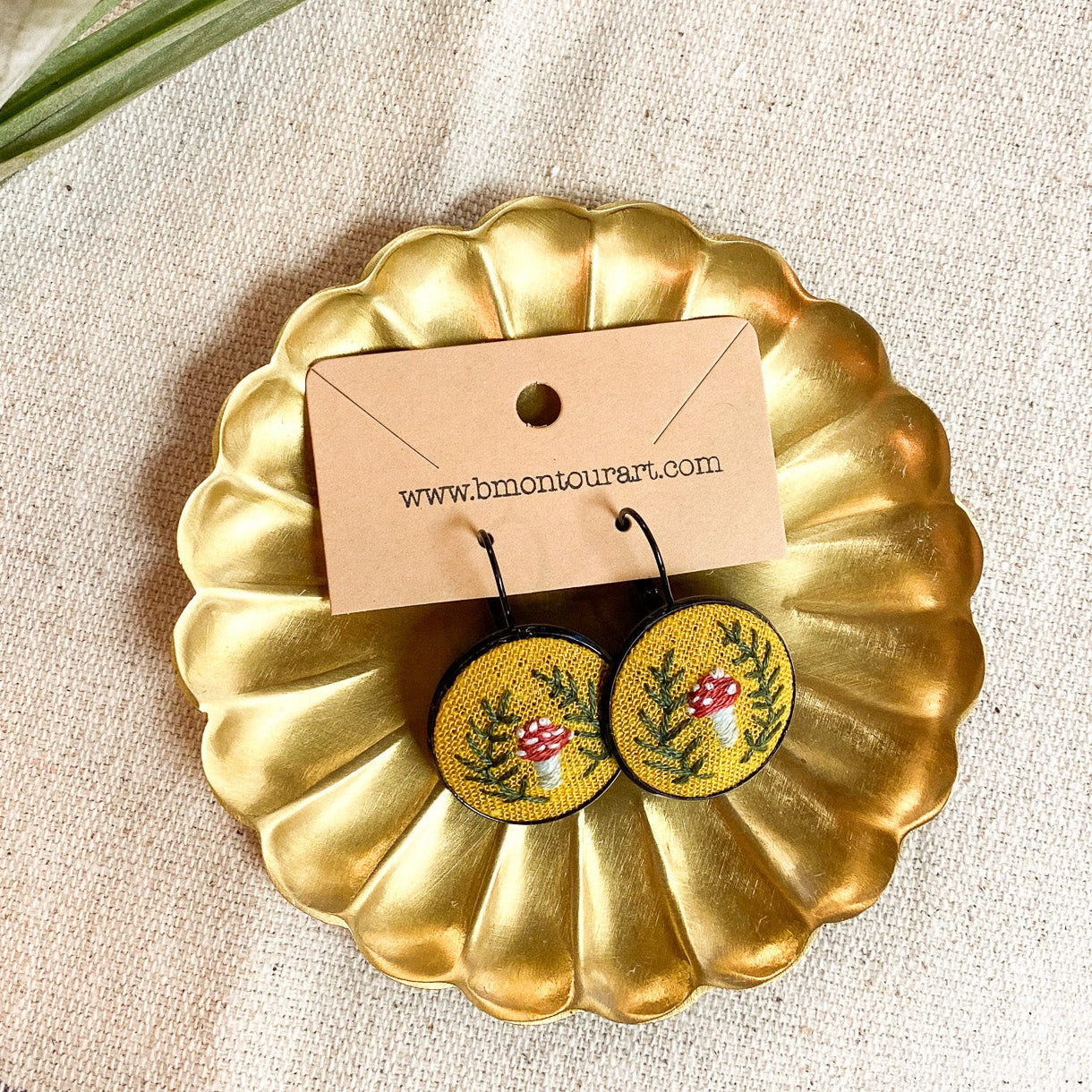 Gold earring dish with floral earrings and a card on a textured surface with leaves.