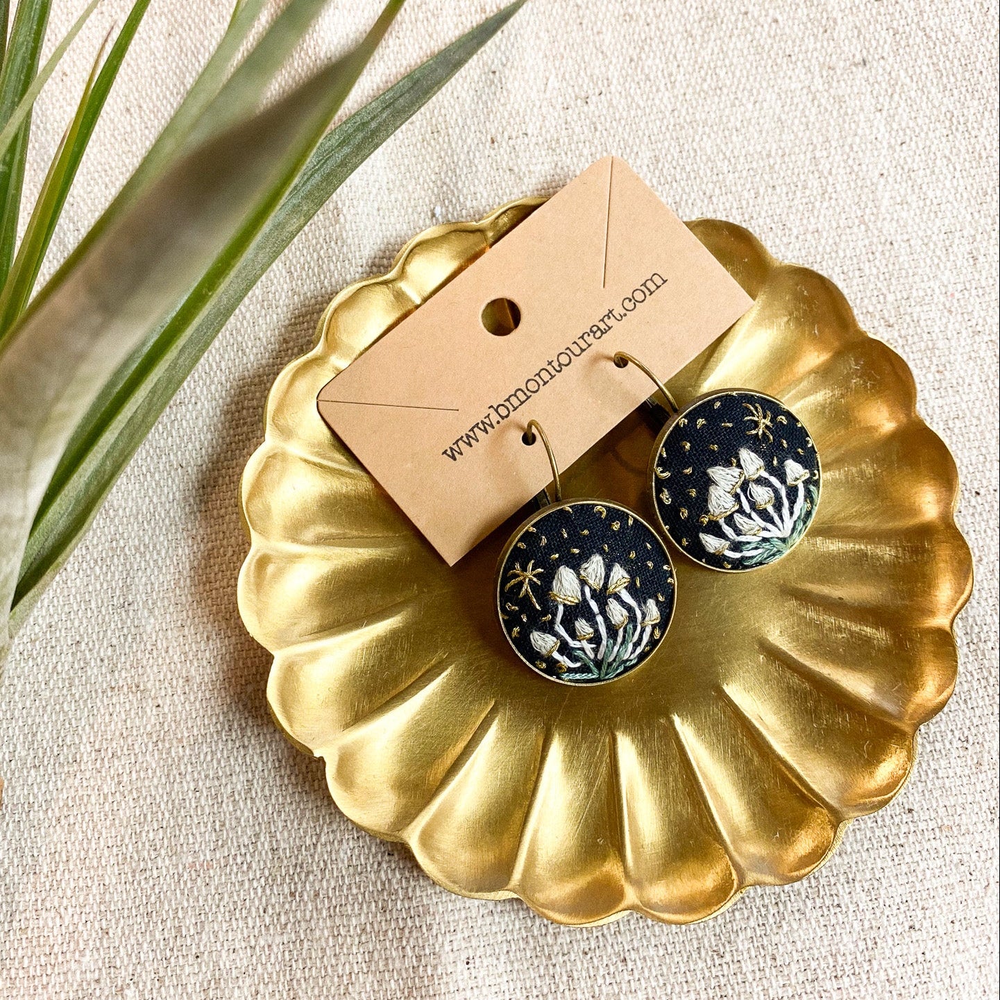 Black floral earrings on a gold dish with a branded card, placed on a textured surface with a plant leaf.
