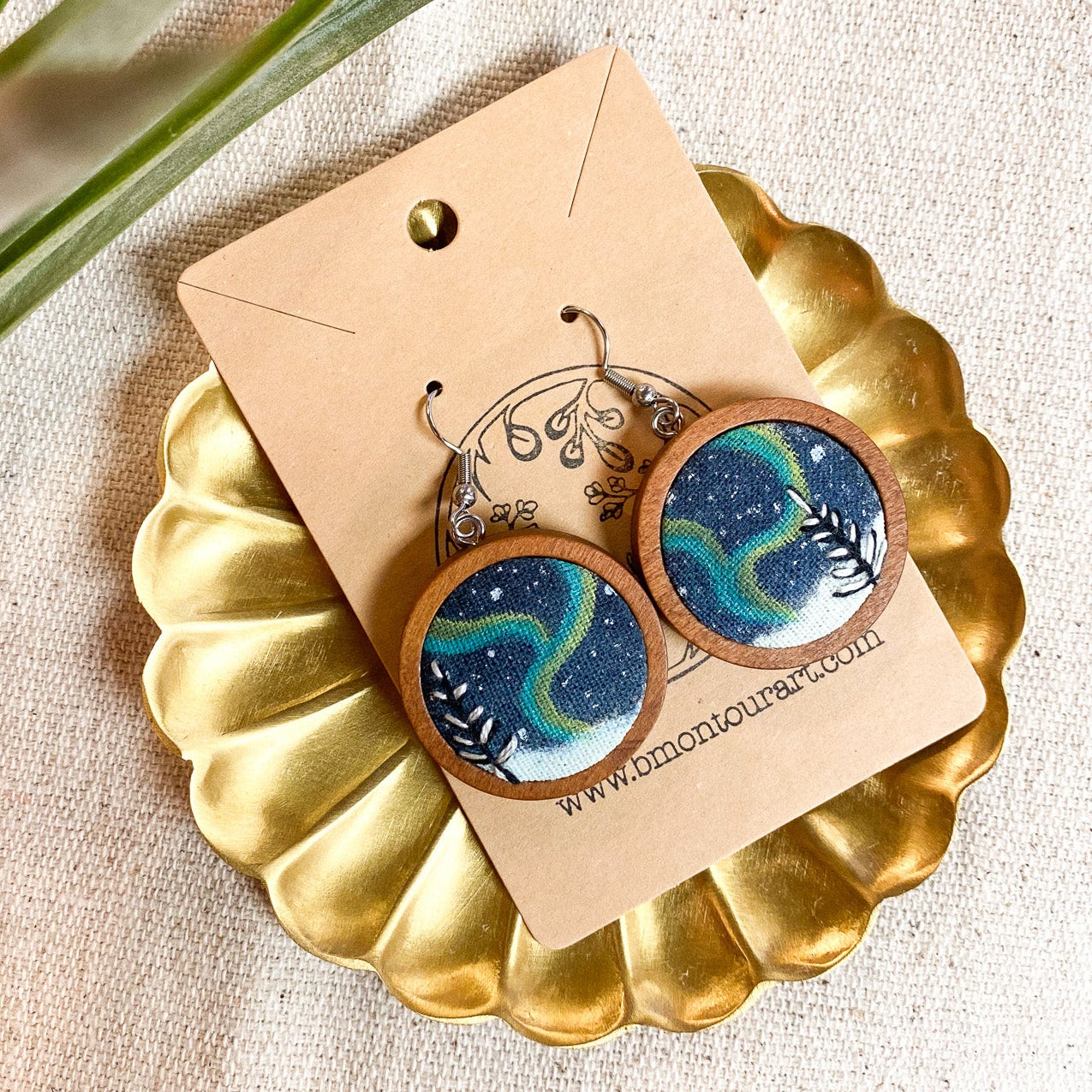 Wooden earrings with blue and green pattern on a gold stand with a beige card, placed on a textured surface with a plant leaf.