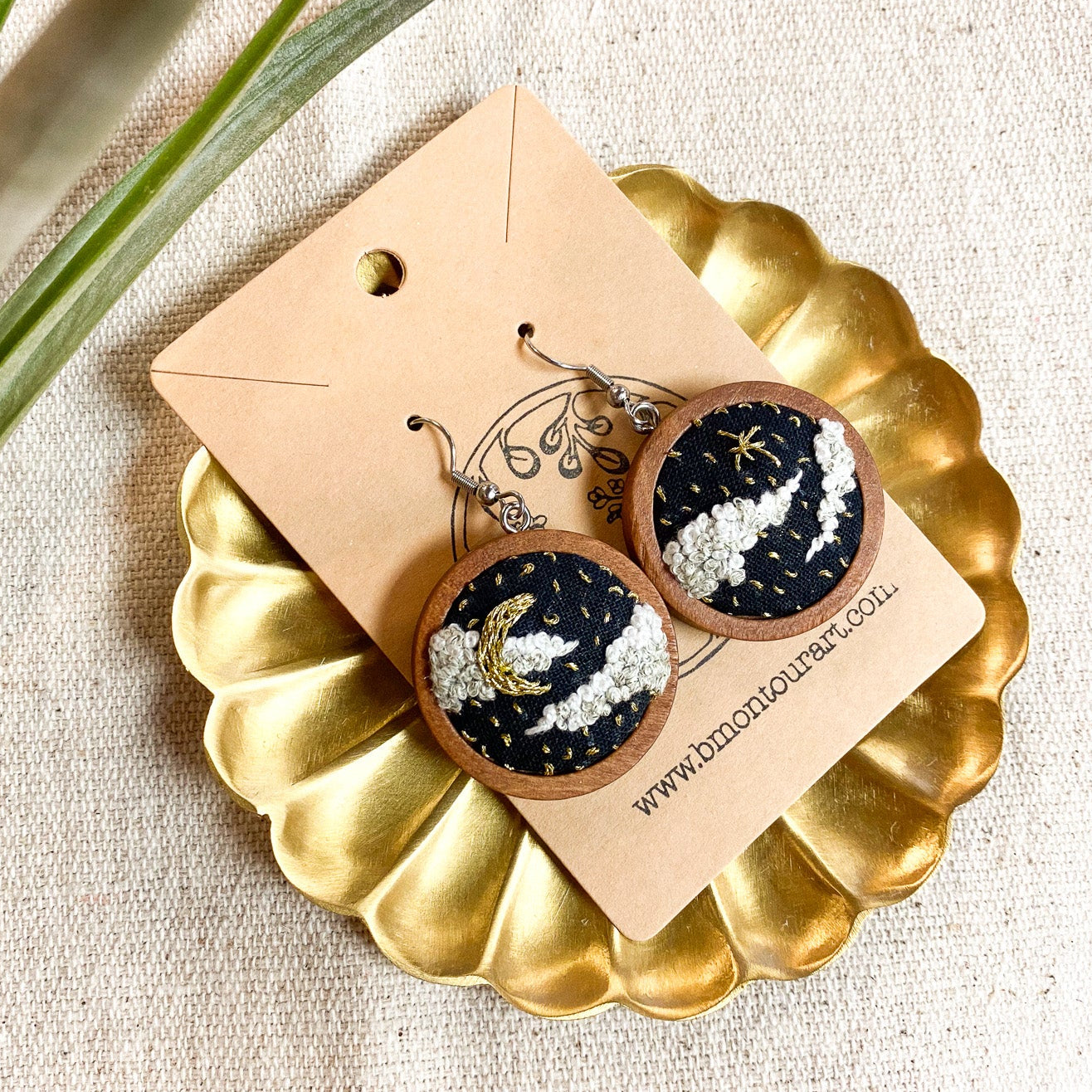 Pair of earrings on a gold dish with a beige card, placed on a textured surface with a plant leaf.