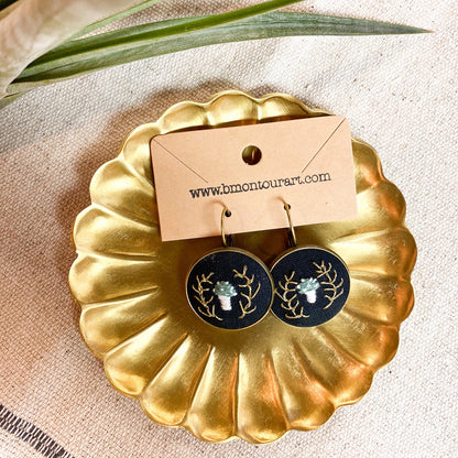Gold-framed earrings with a nature-themed design on a beige surface with a plant leaf.