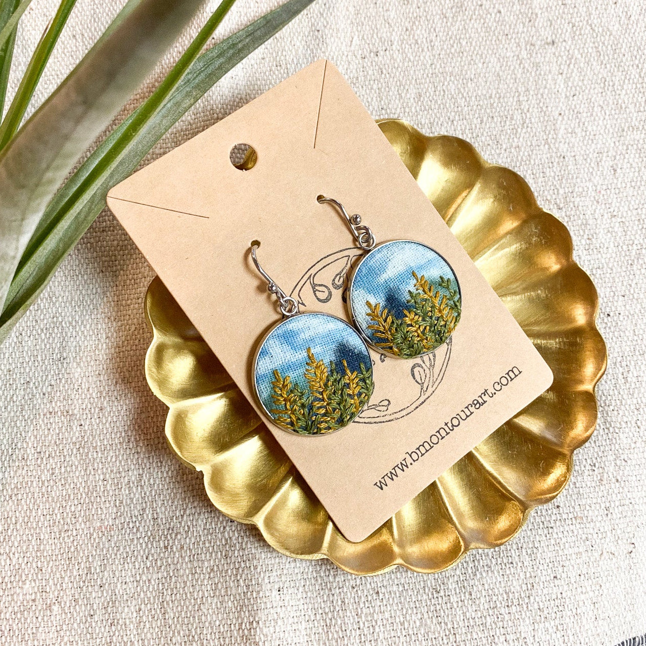 Pair of earrings with nature-themed design on a gold stand with a card, placed on a textured surface.