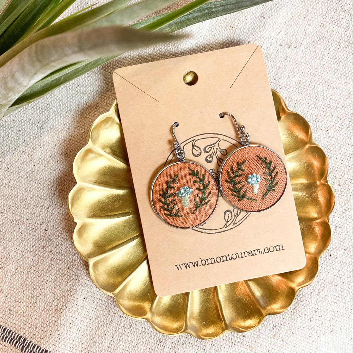 Pair of earrings on a gold stand with a matching card, placed on a textured surface with a plant in the corner.