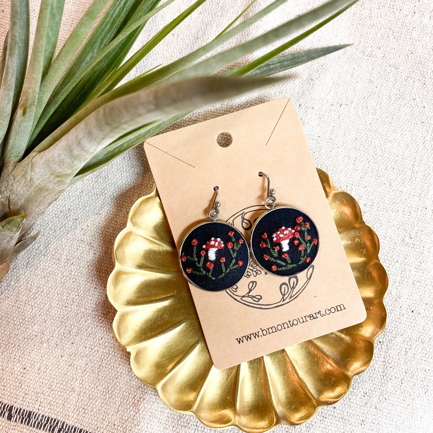 Pair of earrings on a gold stand with a decorative card, surrounded by green leaves.