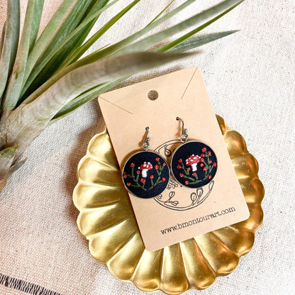 Pair of earrings on a gold stand with a decorative card, surrounded by green leaves.