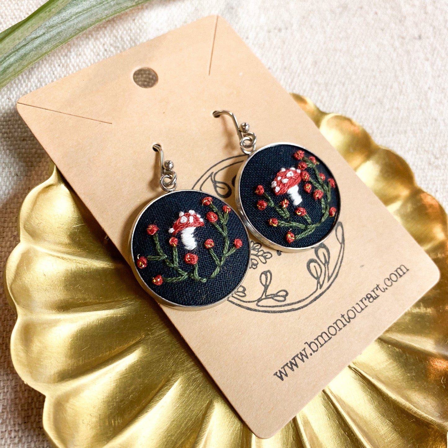Embroidered earrings on a gold stand with a beige card displaying a website URL.