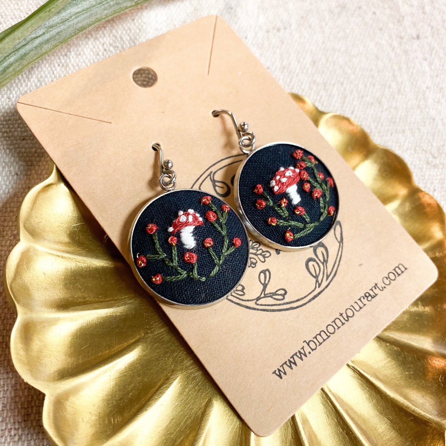 Embroidered earrings on a gold stand with a beige card displaying a website URL.