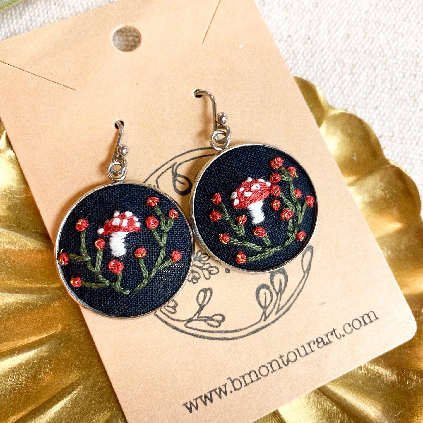 Embroidered earrings with mushroom design on a branded card, placed on a gold surface.