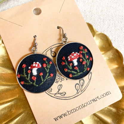 Embroidered earrings with mushroom design on a branded card, placed on a gold surface.