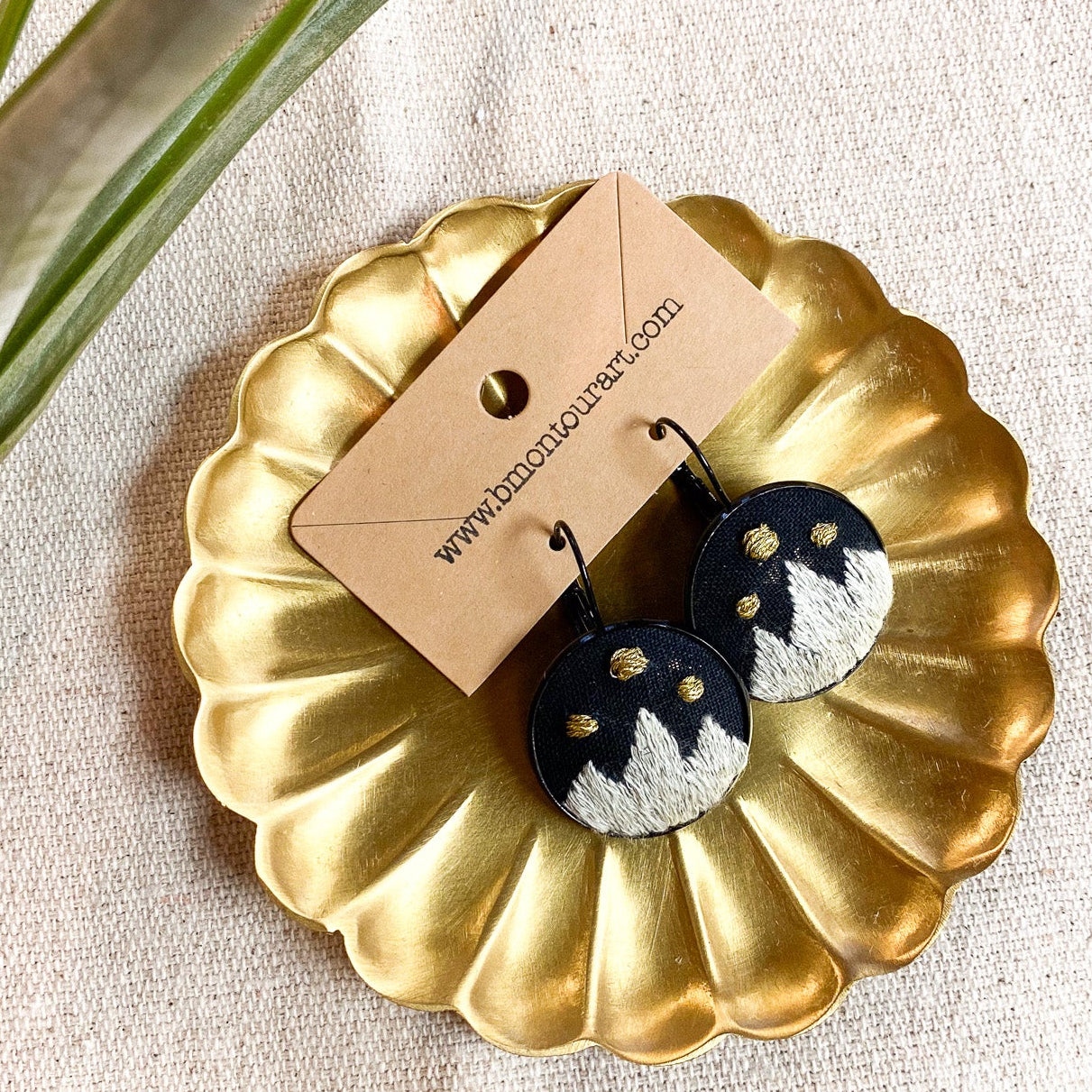 Pair of earrings on a gold dish with a plant in the background