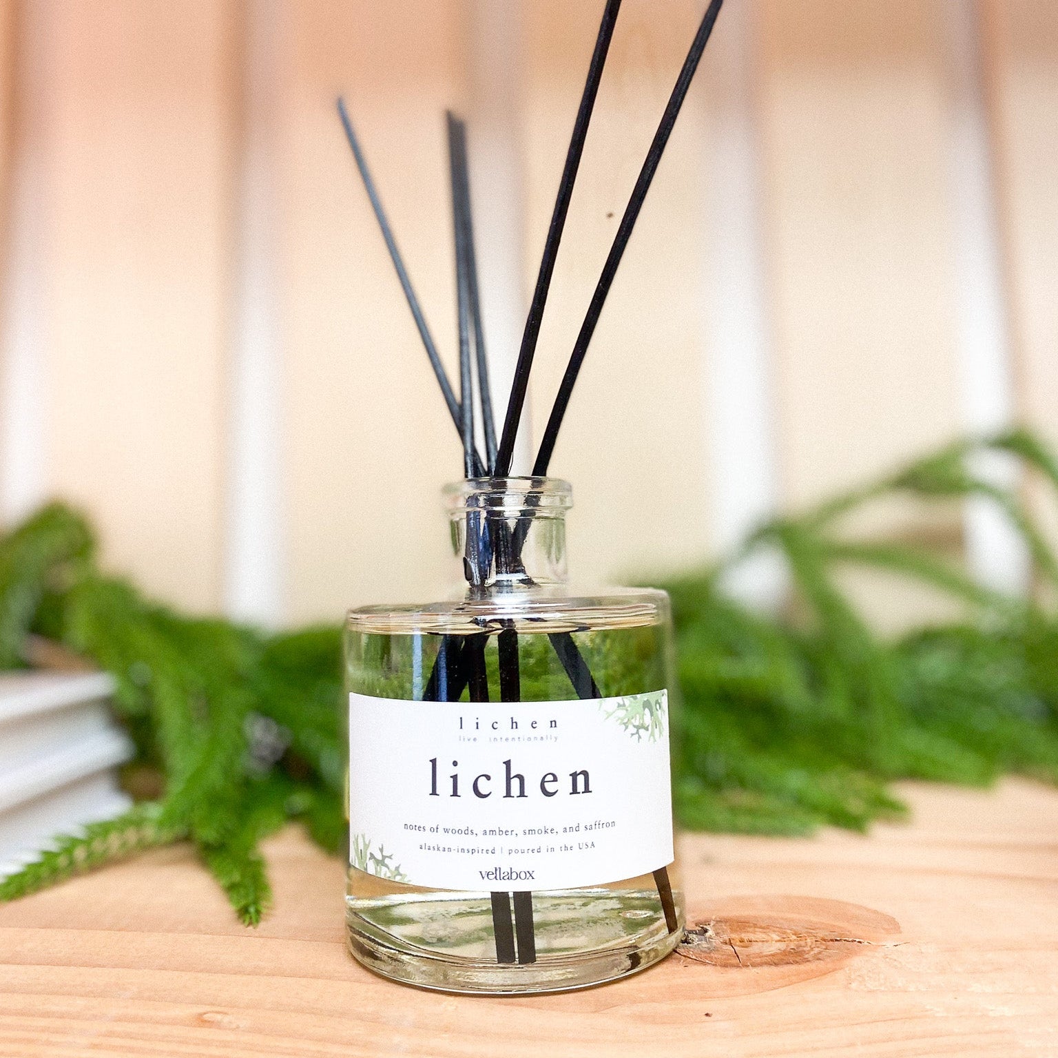 Diffuser bottle labeled 'lichen' with black sticks on a wooden surface with greenery in the background