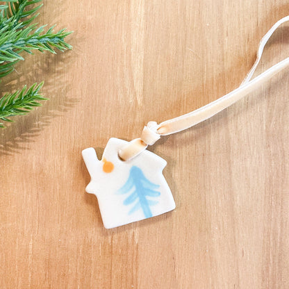 Ceramic ornament with a house and tree design on a wooden surface with greenery.