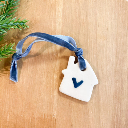 Ceramic white house-shaped ornament with a blue ribbon and a heart on a wooden surface with greenery.