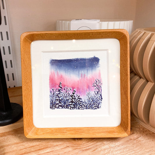 Framed artwork of a snowy landscape with a pink and blue northern lights on a wooden surface.