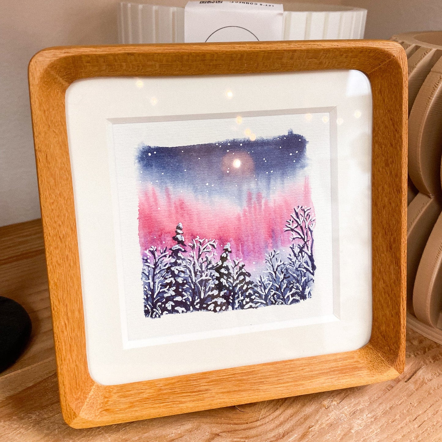 Framed artwork of a snowy landscape with a pink and blue northern lignts water color, placed on a wooden surface.