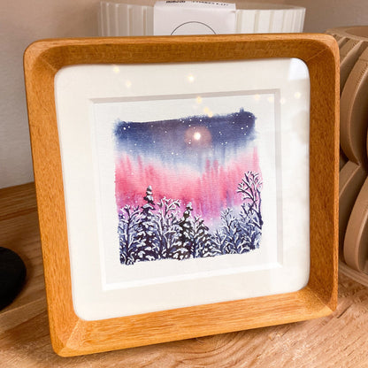 Framed artwork of a snowy landscape with a pink and blue northern lignts water color, placed on a wooden surface.