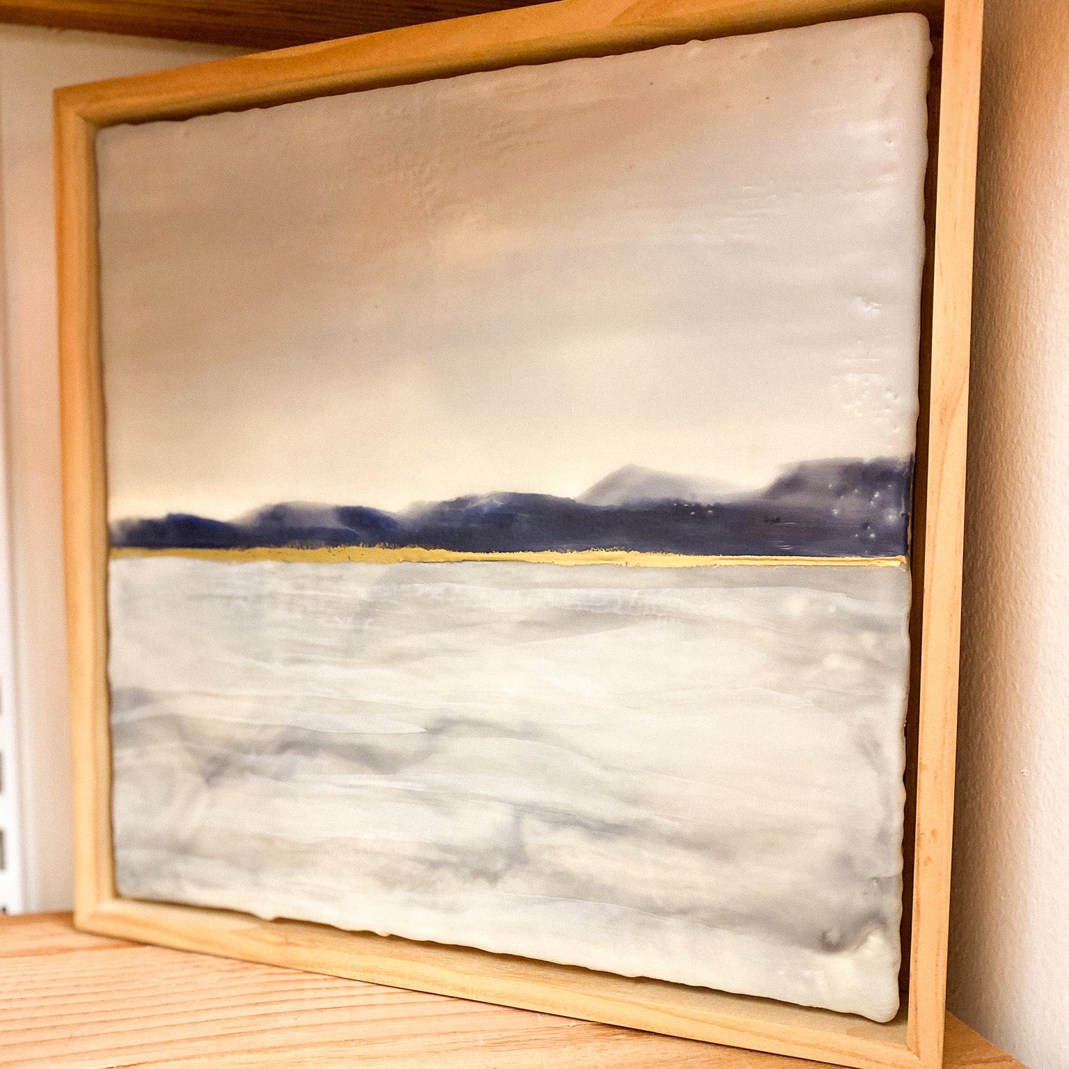 Framed painting of a landscape with mountains on a wooden shelf.