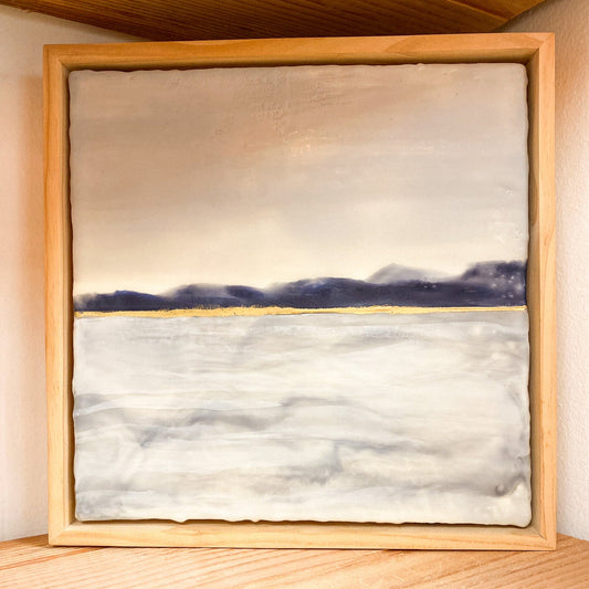Framed artwork of a landscape with water and sky on a wooden surface.