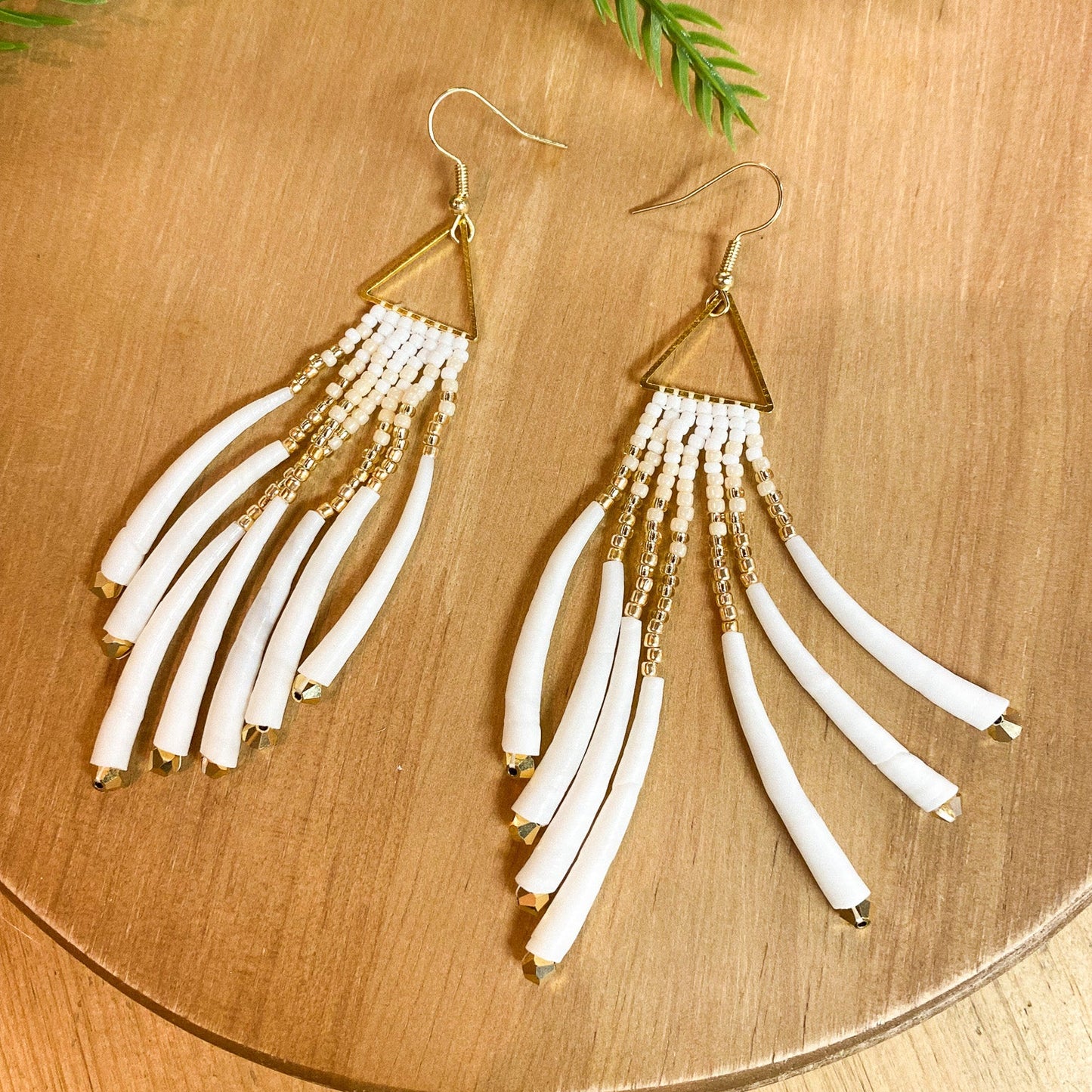 Pair of gold and white fringe earrings on a wooden surface with green leaves.