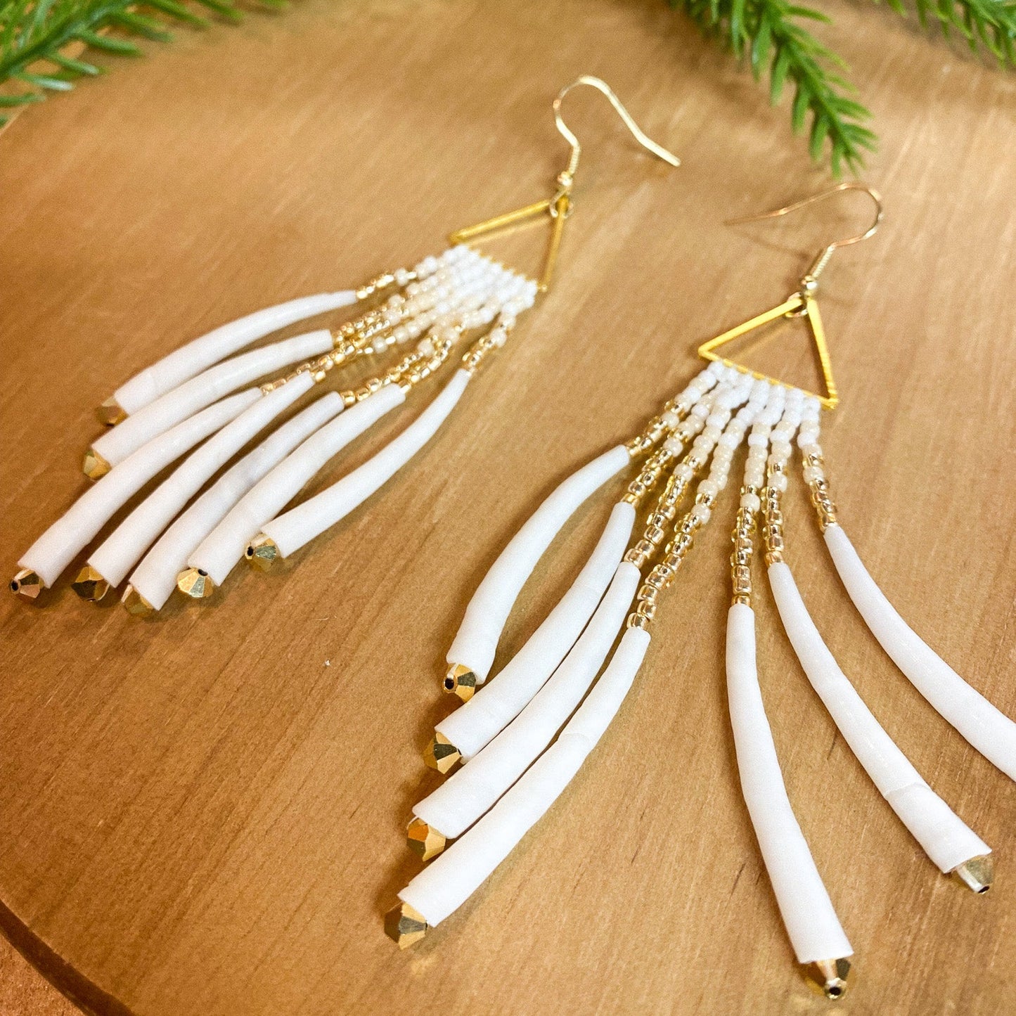Gold and white fringe earrings on a wooden surface with green leaves in the background