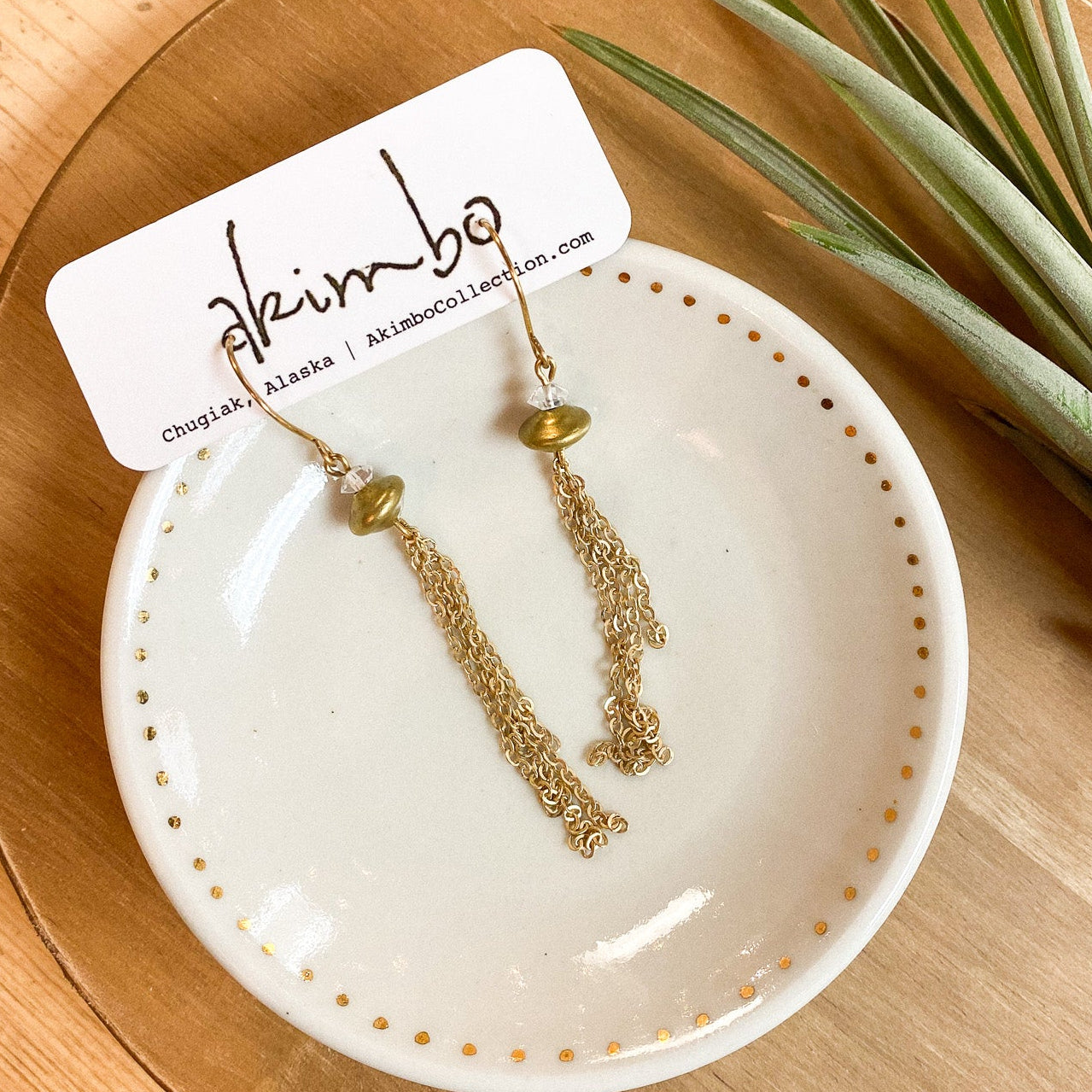 Gold earrings on a white dish with a 'Akimbo' tag, placed on a wooden surface with a plant.