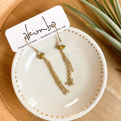 Gold earrings on a white dish with a 'Akimbo' tag, placed on a wooden surface with a plant.