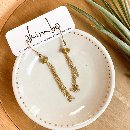 Gold earrings on a white dish with a 'Akimbo' tag, placed on a wooden surface with a plant.