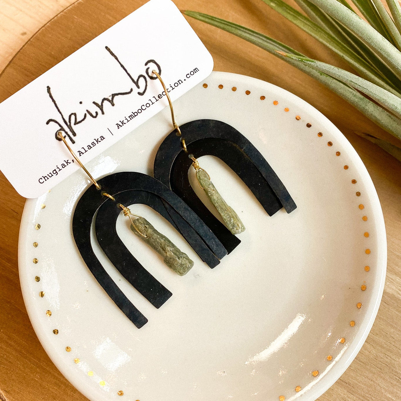Black and green earrings on a white dish with a brand card, on a wooden surface with a plant.
