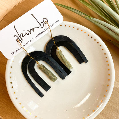 Black and green earrings on a white dish with a brand card, on a wooden surface with a plant.