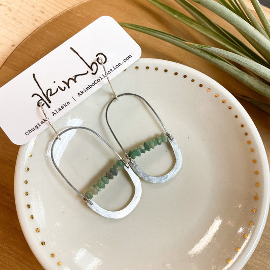 Silver earrings with jade green stones on a white dish with gold dots, branded 'Akimbo'.